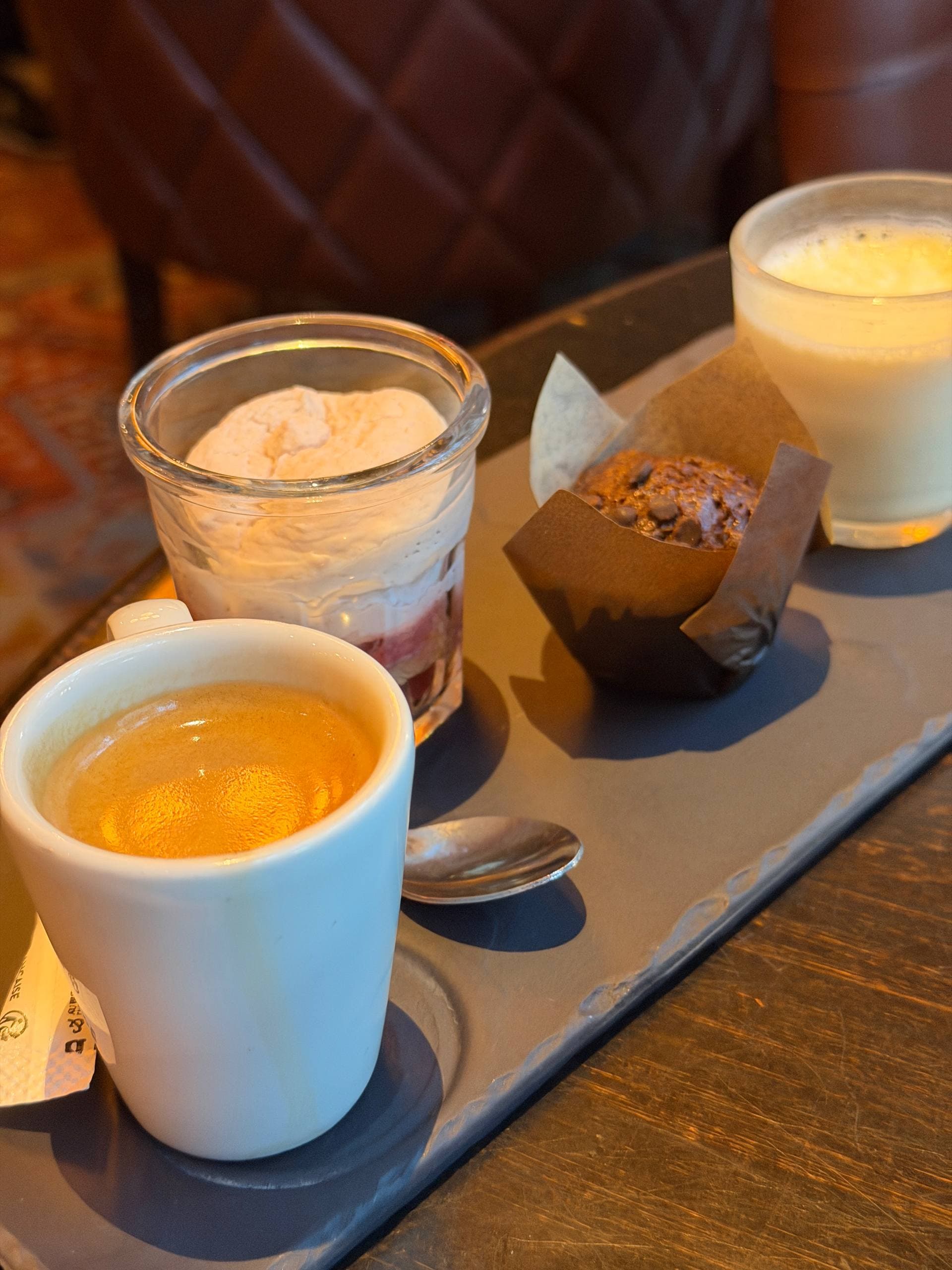Phone photograph of a café gourmand tray with espresso, a cream dessert, a chocolate muffin, and a pale drink